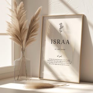 May include: A gold framed print with a white background and black text. The text reads "ISRAA" with the date "17/03/2025" below. The Arabic word for "Israa" is written in black below the date. The text below the Arabic word explains the meaning of "Israa" in Islam. The print is sitting on a white surface with a vase of dried pampas grass in front of it.