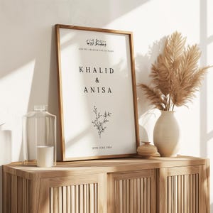 Peut inclure: Une impression encadr&eacute;e blanche avec du texte noir qui dit "Et Nous Vous Avons Cr&eacute;&eacute;s Par Paires" en &eacute;criture arabe, suivie de la traduction anglaise. Sous le texte figurent les noms "Khalid & Anisa" et la date "16 juin 2024". Une petite illustration florale est centr&eacute;e sous les noms.