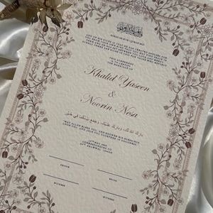 Puede incluir: Un certificado de boda con borde floral y texto en ingl&eacute;s y &aacute;rabe. Los nombres "Khalid Yaseen & Noorin Nesa" se destacan. Flores secas se colocan cerca del certificado. El certificado est&aacute; sobre un fondo de sat&eacute;n blanco.