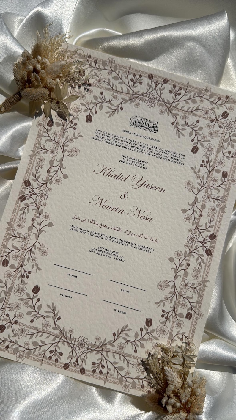 Puede incluir: Un certificado de boda con borde floral y texto en ingl&eacute;s y &aacute;rabe. Los nombres "Khalid Yaseen & Noorin Nesa" se destacan. Flores secas se colocan cerca del certificado. El certificado est&aacute; sobre un fondo de sat&eacute;n blanco.