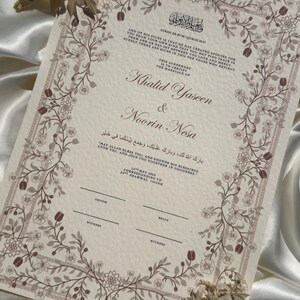 Puede incluir: Un certificado de boda con borde floral y texto. Los nombres "Khalid Yaseen & Noorin Nesa" se muestran de forma destacada. El certificado incluye escritura &aacute;rabe y texto en ingl&eacute;s. Flores secas se colocan cerca del certificado.