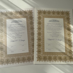 Peut inclure: Deux certificats blancs avec des bordures orn&eacute;es dor&eacute;es et du texte arabe. Les certificats sont pour une c&eacute;r&eacute;monie de mariage et sont dat&eacute;s de d&eacute;cembre 2021. Le texte en bas indique "Bilal & Amaya".
