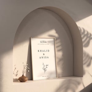 Peut inclure: Une impression encadr&eacute;e avec un fond blanc et du texte noir. Le texte dit "Et nous vous avons cr&eacute;&eacute;s en paires" en &eacute;criture arabe, suivi de la traduction anglaise. Sous le texte figurent les noms "Khalid & Anisa" et la date "16 juin 2024". Une petite illustration florale est centr&eacute;e sous les noms.