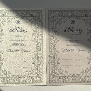 Op de afbeelding: Twee ivoorwitte huwelijkscertificaten met sierlijke bloemranden en Arabische kalligrafie. De tekst luidt "En Wij hebben jullie in paren geschapen" en "Deze overeenkomst is gesloten op 18 mei 2021, overeenkomend met 14 Shawwal 1442 AH, om het huwelijk van Khalid en Jasmine te bevestigen".