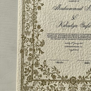 K&ouml;nnte beinhalten: Eine cremefarbene Heiratsurkunde mit einem goldenen floralen Rand. Der Text lautet: "Diese Vereinbarung dient dazu zu bescheinigen, dass die Ehe von Muhammad A & Kohadija Safa in &Uuml;bereinstimmung mit den Anforderungen des islamischen Rechts geschlossen wurde, wie es im Heiligen Koran und den Traditionen des Propheten Muhammad (SAW) verankert ist, mit den vereinbarten Zeugen in Anwesenheit des Standesbeamten und der Zeugen. Samstag, 13. Januar 2024, entsprechend dem 1. Rajab 1445ah."