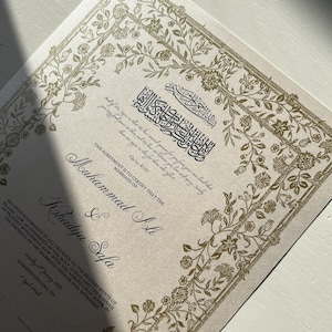 May include: A gold-embossed Islamic marriage certificate with ornate floral borders. The text reads "This agreement is to certify that the marriage of Muhammad Ali & Khadija Safa has been solemnised in accordance to the requirements of Islamic law, as enshrined in the Holy Quran and the Sunnah of Prophet Muhammad (SAW) with the agreed upon mahr, in the presence of the marriage officer and witnesses."