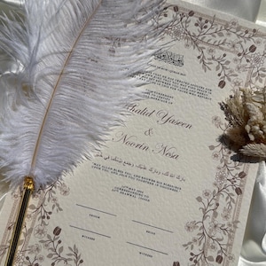 Puede incluir: Una invitaci&oacute;n de boda con borde floral y los nombres "Khalid Yaseen & Noorin Nesa" en escritura elegante. Una pluma blanca con punta dorada descansa sobre la invitaci&oacute;n. El fondo es de color crema.
