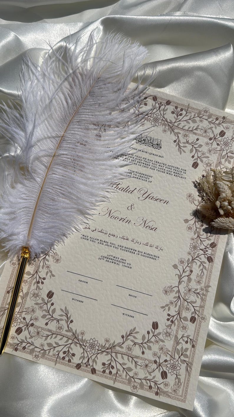 Puede incluir: Una invitaci&oacute;n de boda con borde floral y los nombres "Khalid Yaseen & Noorin Nesa" en escritura elegante. Una pluma blanca con punta dorada descansa sobre la invitaci&oacute;n. El fondo es de color crema.