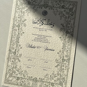 Op de afbeelding: Een wit certificaat met een zwarte bloemmotiefrand en tekst. De tekst luidt "En Wij hebben jullie in paren geschapen, Koran 78:8." Het certificaat is voor het huwelijk van Khalid en Jasmine. De datum van de overeenkomst is 14 mei 2023, overeenkomend met 24 Shawwal 1444 AH.