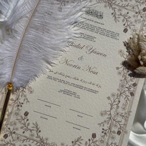 Puede incluir: Un certificado de boda con borde floral y caligraf&iacute;a, con los nombres "Khalid Yaseen & Noorin Nesa." Una pluma blanca descansa sobre el documento, sobre un fondo de sat&eacute;n.