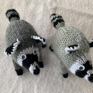 May include: Two handmade knitted raccoon toys featuring grey bodies, black and white facial markings, striped tails, and plastic googly eyes. Each plush animal is crafted with a textured yarn finish, pointed ears, and small black paws, presented from a top-down perspective.