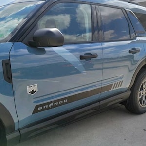 May include: A blue Ford Bronco Sport with black trim and a black and silver stripe along the side. The vehicle has a "Trail Boss" badge on the side and the word "Bronco" in black lettering on the side.