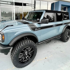 May include: A light blue Ford Bronco with black accents and black wheels. The vehicle has a black roof and a black step bar. The word "Bronco" is visible on the side of the vehicle.