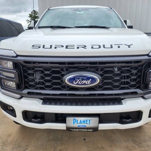 Könnte beinhalten: Ein weißer Ford Super Duty Pickup mit einem schwarzen Kühlergrill und einem schwarzen Ford-Logo. Der Pickup hat einen schwarzen Schriftzug "SUPER DUTY" auf der Motorhaube und ein Planet Ford Händlerlogo auf der vorderen Stoßstange.