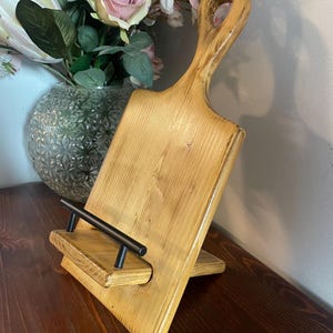May include: A wooden phone stand with a rustic finish. The stand is made from a repurposed cutting board with a handle. It has a black metal phone holder.