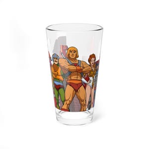 May include: A clear glass featuring a colorful illustration of He-Man and the Masters of the Universe characters. The design includes He-Man, Teela, Man-At-Arms, and other heroes. The glass is a standard pint size.