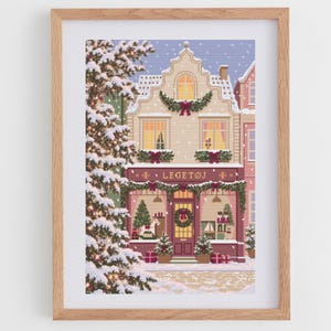 May include: A framed cross-stitch art piece depicting a snowy winter scene of a toy store labeled LEGETØJ. The building features festive greenery, red bows, and warm glowing windows. A large snow-covered pine tree stands to the left of the shop.