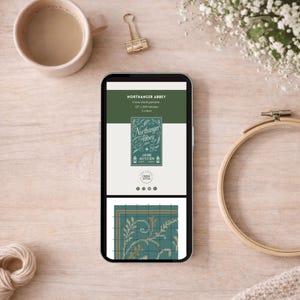 May include: A smartphone displays a cross-stitch pattern for "Northanger Abbey" by Jane Austen. The screen shows a teal and gold design, with the book title and author's name. A cup of coffee, a gold binder clip, and a wooden embroidery hoop are also visible.
