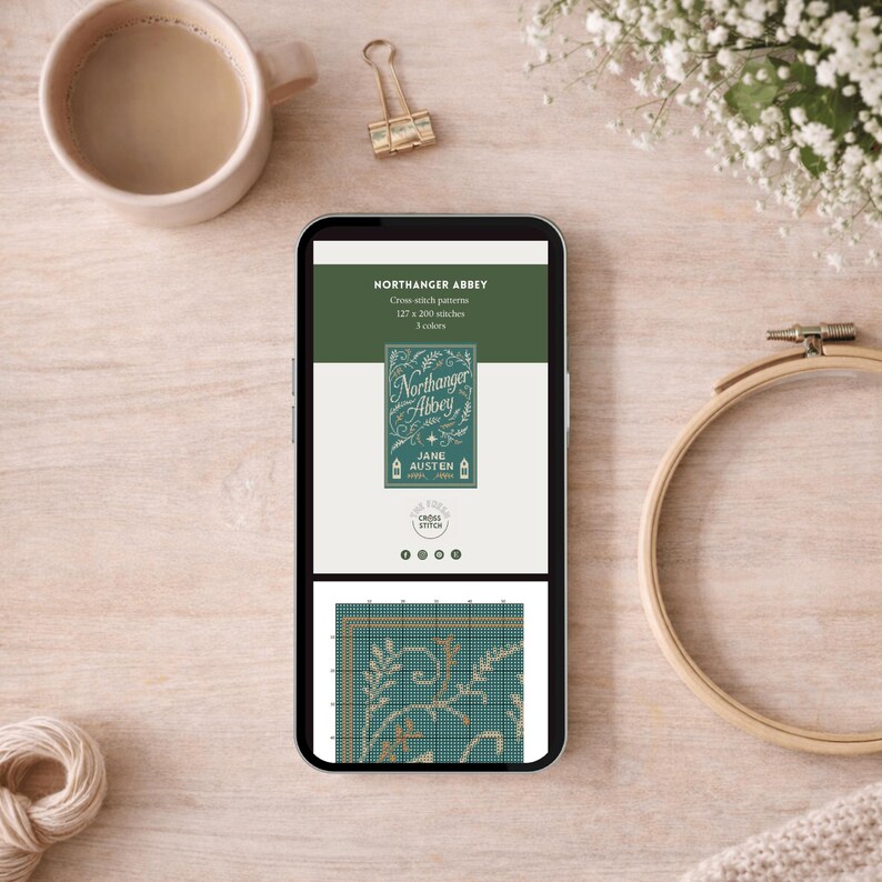 May include: A smartphone displays a cross-stitch pattern for "Northanger Abbey" by Jane Austen. The screen shows a teal and gold design, with the book title and author's name. A cup of coffee, a gold binder clip, and a wooden embroidery hoop are also visible.