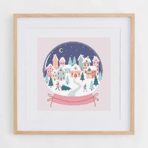 May include: A framed cross-stitch pattern depicting a winter snow globe scene with a small village, snow-covered houses, evergreen trees, and two figures carrying a tree, all set against a dark blue night sky with a crescent moon.