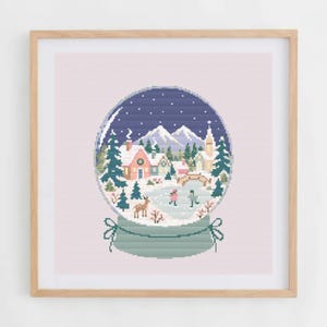 May include: A square framed cross-stitch embroidery depicting a winter snow globe scene. Inside the globe are snow-covered mountains, pine trees, houses, a bridge, a deer, and two figures ice skating on a frozen pond under a dark blue sky.