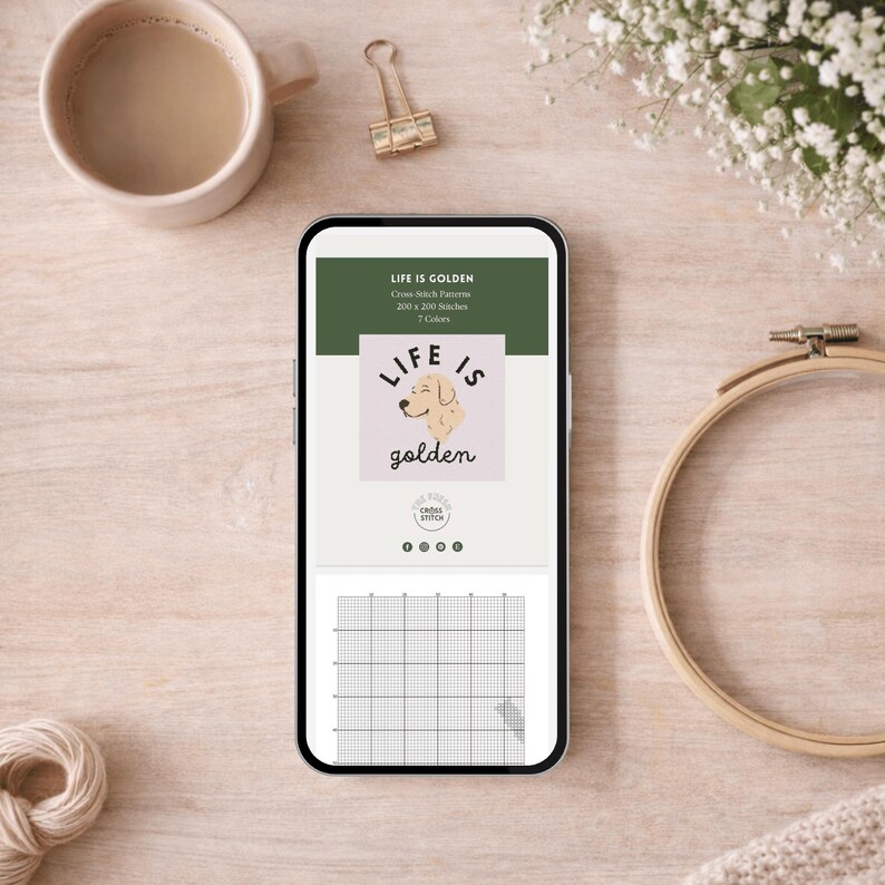 May include: A smartphone displays a cross-stitch pattern with the text "LIFE IS GOLDEN" and a dog illustration. The phone is on a wooden surface, next to a cup of coffee, a gold binder clip, and an embroidery hoop.