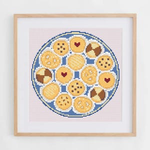 May include: A framed cross-stitch embroidery featuring a circular blue platter filled with various golden-brown cookies. The cookies include chocolate chip, heart-centered, and checkerboard patterns, all rendered in detailed needlework stitches.