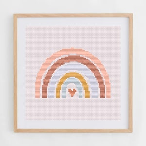May include: A framed cross-stitch artwork featuring a rainbow design. The rainbow has layers of peach, brown, light blue, yellow, and a heart in the center. The background is a light pink color, and the frame is made of light-colored wood.