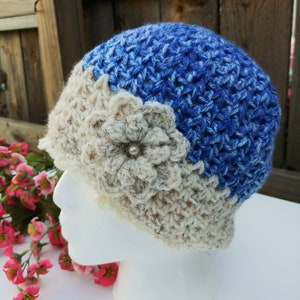 May include: A handmade crochet hat in blue and cream colors. The hat features a floral embellishment with a pearl center. The hat is displayed on a white mannequin head, showcasing the intricate stitch work and design.