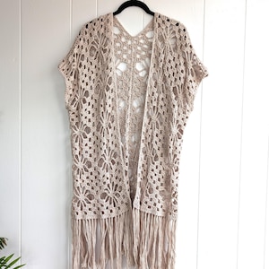 May include: A beige, open-front crochet cardigan with short sleeves and a fringed hem. The intricate floral pattern is visible throughout the garment. The cardigan is hanging on a black hanger against a white wall.