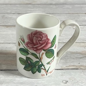 Può includere: Tazza in ceramica bianca con un'illustrazione di una rosa rosa, foglie verdi e boccioli. La tazza ha un manico curvo. Le parole "ROSA Royal" e "MAJESTAS" sono stampate sul lato.