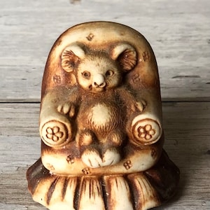 May include: A ceramic figurine of a brown koala bear sitting in a brown armchair with floral details. The figurine is a light brown color with brown accents.