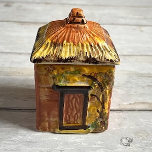 Op de afbeelding: Een keramische suikerpot in de vorm van een huisje met een rieten dak, bruine bakstenen muren, een gele deur en groene wijnstokken. De pot is versierd met gele en bruine verf.