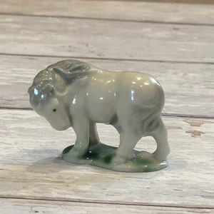 May include: A white porcelain figurine of a cow with a green base. The cow is standing with its head down, as if it is grazing.