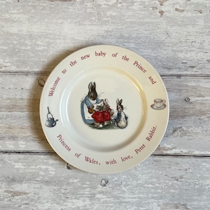 May include: A white ceramic plate with a red border and a picture of three bunnies. The plate has the text "Welcome to the new baby of the Prince and Princess of Wales, with love, Peter Rabbit."