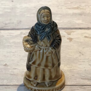 May include: A ceramic figurine of a woman wearing a brown dress and a blue shawl, holding a basket. The figurine is standing on a base with the word "KATHLEEN" written on it.