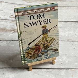 Op de afbeelding: Een vintage Ladybird Children's Classics boek getiteld "Tom Sawyer" met een kleurrijke illustratie van twee jongens op een vlot in een rivier. Het boek wordt getoond op een klein houten ezel.
