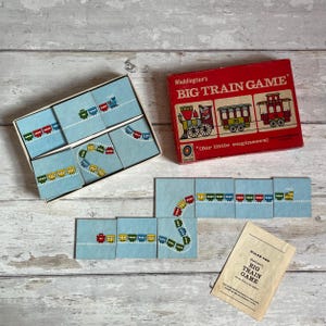 May include: A vintage "Big Train Game" by Waddington's. The red box features train illustrations. The game includes blue tiles with colorful train cars and a booklet with rules. The game is designed for little engineers.
