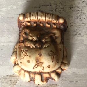 May include: A ceramic figurine of two koalas sleeping in a bed with a floral patterned blanket. The figurine is brown and white.