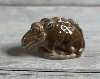 May include: A small, brown ceramic figurine of a dinosaur with a toothy grin. The dinosaur has a textured surface and a slightly raised eye.