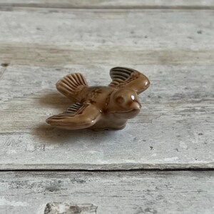 May include: A small, brown ceramic figurine of a bird in flight. The bird has brown and white wings and a brown body.