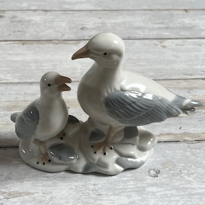 Op de afbeelding: Een wit porseleinen beeldje van twee meeuwen die op een grijze en witte basis zitten. De grotere meeuw heeft grijze vleugels en een wit lichaam, terwijl de kleinere meeuw helemaal wit is.