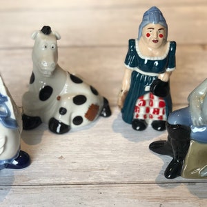 Può includere: Quattro figurine in ceramica di un'oca, una mucca, una donna e un gatto. L'oca indossa un berretto blu e una sciarpa a strisce rosse e bianche. La mucca è bianca e nera con una macchia marrone. La donna indossa un vestito blu con una gonna a quadri rossi e bianchi. Il gatto indossa pantaloni blu e un gilet marrone ed è seduto su una scatola marrone.