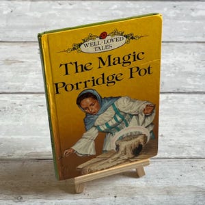 Può includere: Un libro giallo con la copertina verde intitolato "The Magic Porridge Pot" della serie "Well-Loved Tales". La copertina presenta un'illustrazione di una donna con un vestito blu e bianco che versa la pappa da una pentola.