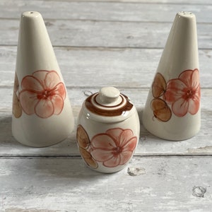 Op de afbeelding: Een set van drie keramische zout- en peperstrooiers en een kruidenpot. De strooiers zijn wit met bruine en roze bloemmotieven. De pot is bruin met een wit bloemmotief.