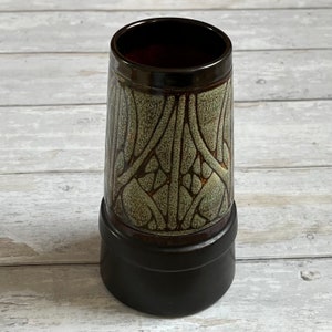 Può includere: Un vaso in ceramica con una smaltatura marrone e verde. Il vaso ha un motivo geometrico e una base larga.