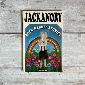 Może przedstawiać: Vintage okładka książki "Jackanory Brer Rabbit Stories". Ilustracja przedstawia królika z kreskówki w ogrodniczkach, na tle tęczy i krajobrazu kwiatowego. Tytuł "Jackanory" jest wyraźnie widoczny.