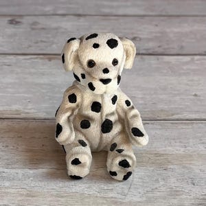 Op de afbeelding: Een kleine, zittende Dalmatische hond figuur met een witte vacht en zwarte vlekken. De hond heeft een vriendelijke uitdrukking met donkere ogen en een zwarte neus. Het beeldje is gemaakt van een textuurrijk materiaal en staat op een houten oppervlak.
