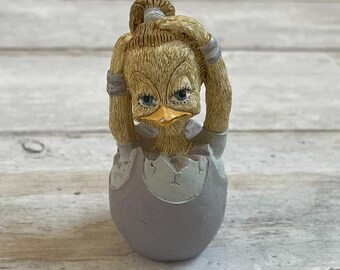 Peut inclure: Une petite figurine en céramique d'un poussin jaune avec une queue de cheval, qui sort d'un œuf gris fissuré. Le poussin a un bec jaune et des yeux bleus.