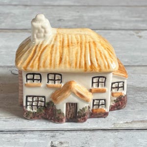 Puede incluir: Figurita de cerámica de una casa de campo con tejado de paja y varias ventanas. La casa está pintada de blanco con detalles marrones y beige, incluyendo el tejado, la puerta y los marcos de las ventanas. Pequeñas plantas verdes y marrones en la base.